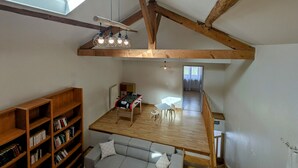 Ein Loft-Zimmer mit schrägem Dach, freiliegenden Holzbalken, Dachfenster, einem Kicker, einer Couch und einem Bücherregal.