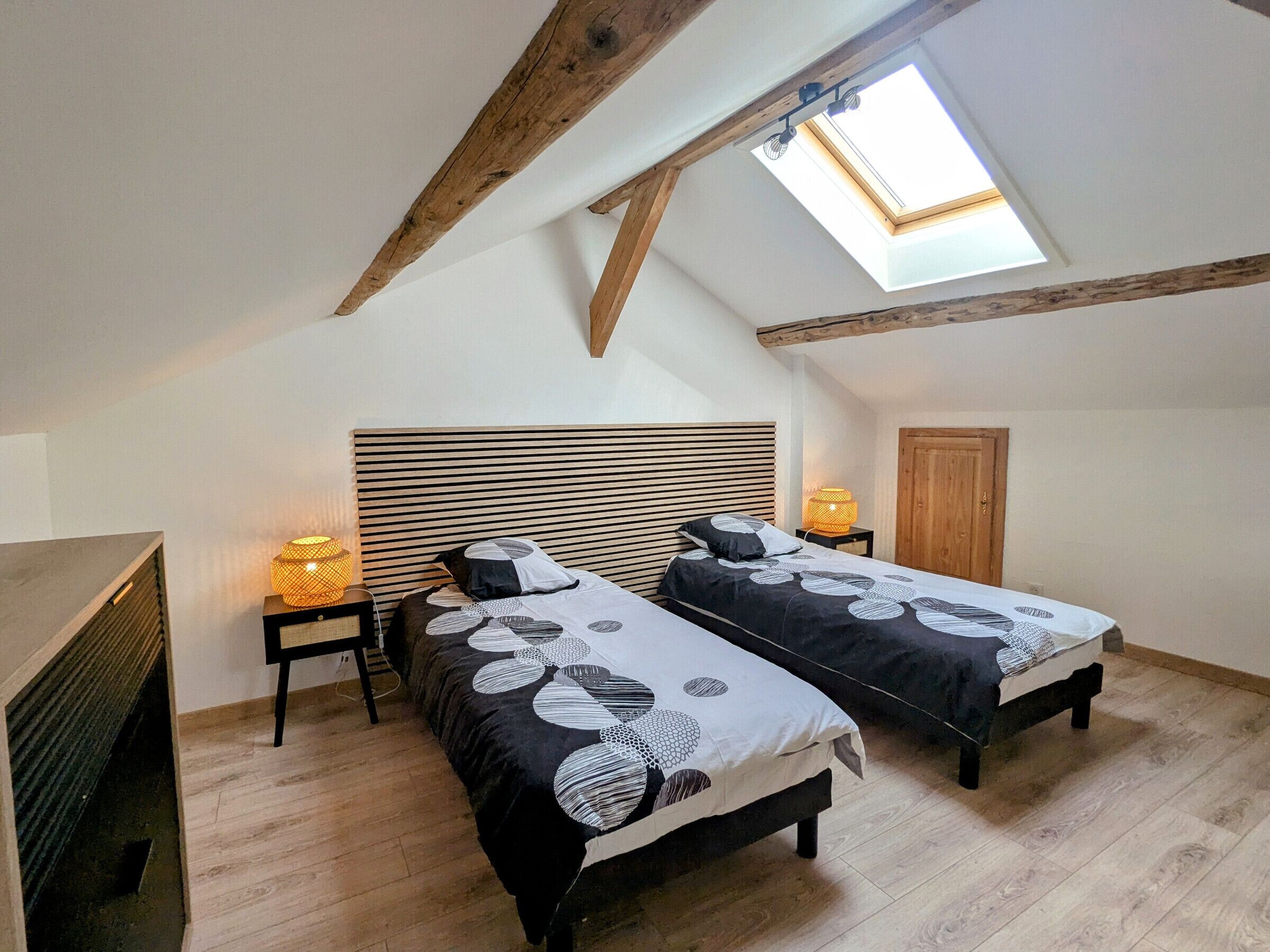 A bedroom with two beds, a skylight, wooden beams, and a nightstand with a lamp.