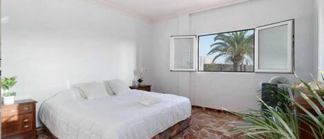 A bedroom with a bed, a nightstand, a potted plant, and a window with a view of palm trees.