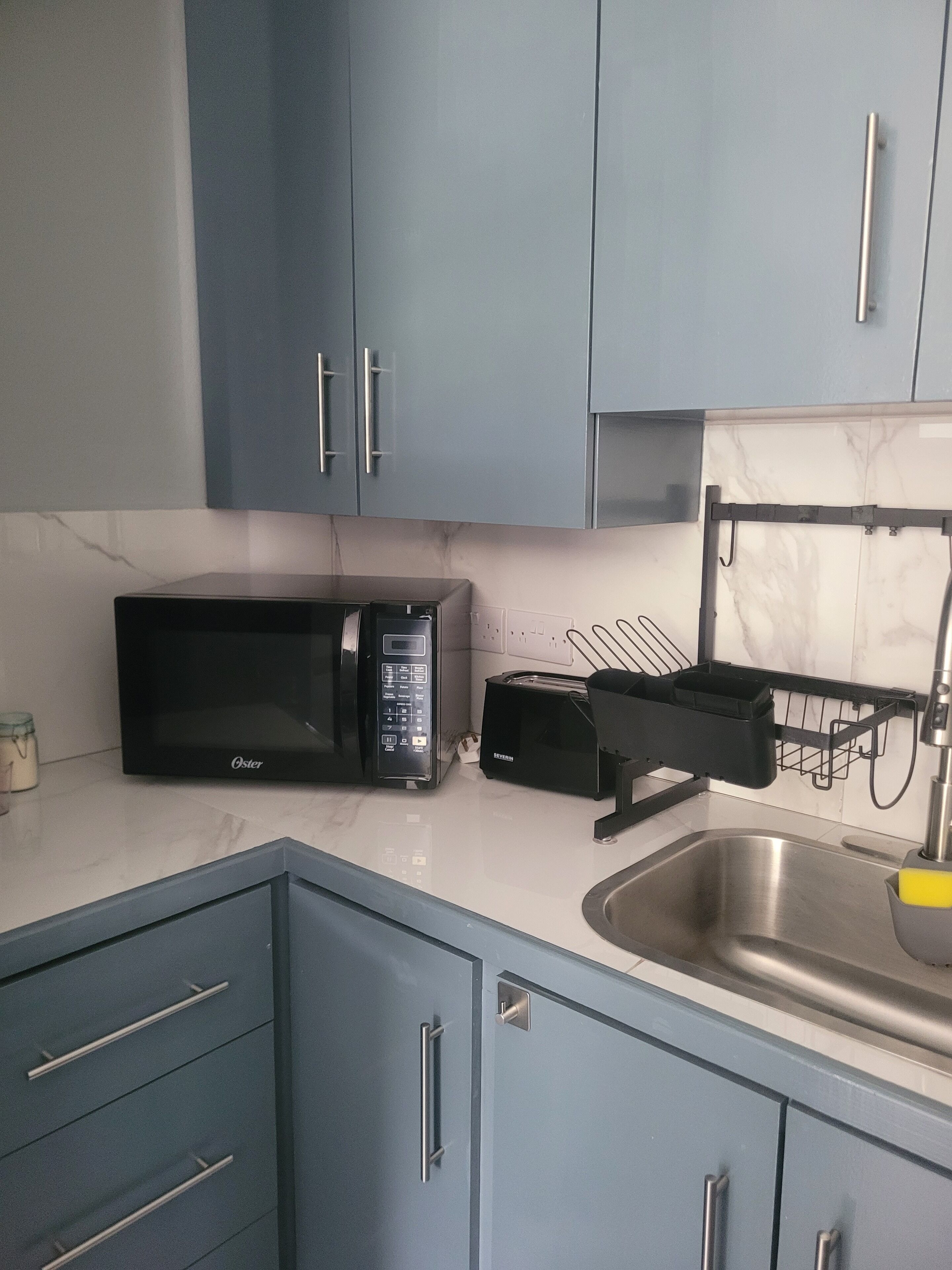 Una cocina con armarios azules, un microondas Oster negro, un tostador y un lavamanos de acero inoxidable.