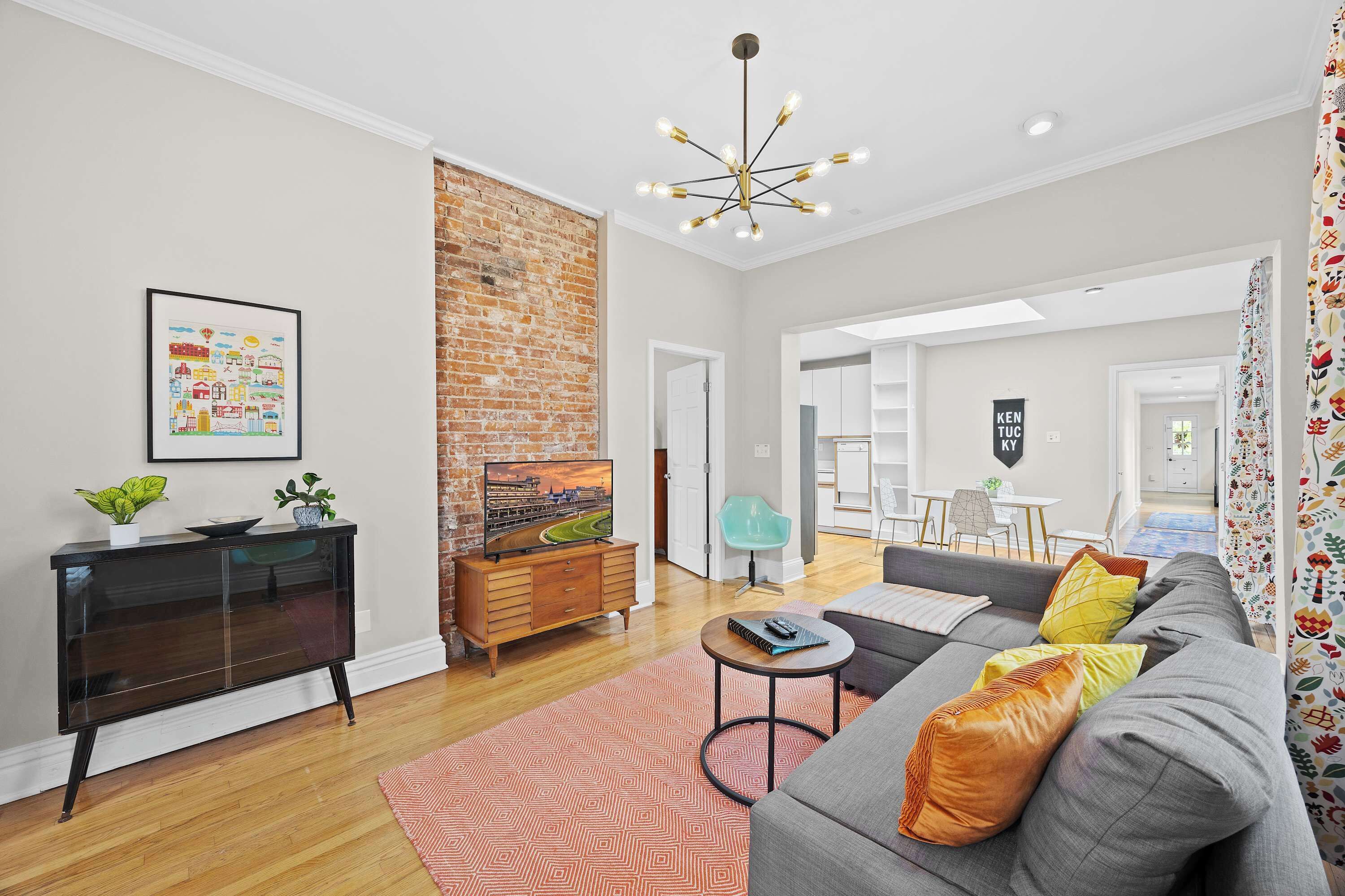 A modern living room with a sofa, a coffee table, a TV on a wooden cabinet, a brick wall, and a ceiling light fixture.