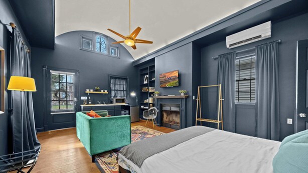 A modern bedroom with a large bed, a ceiling fan, a fireplace, and a view of the outdoors through a window.