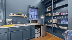 A compact kitchen with a microwave, sink, and a small refrigerator. There are shelves with books and decorative items.