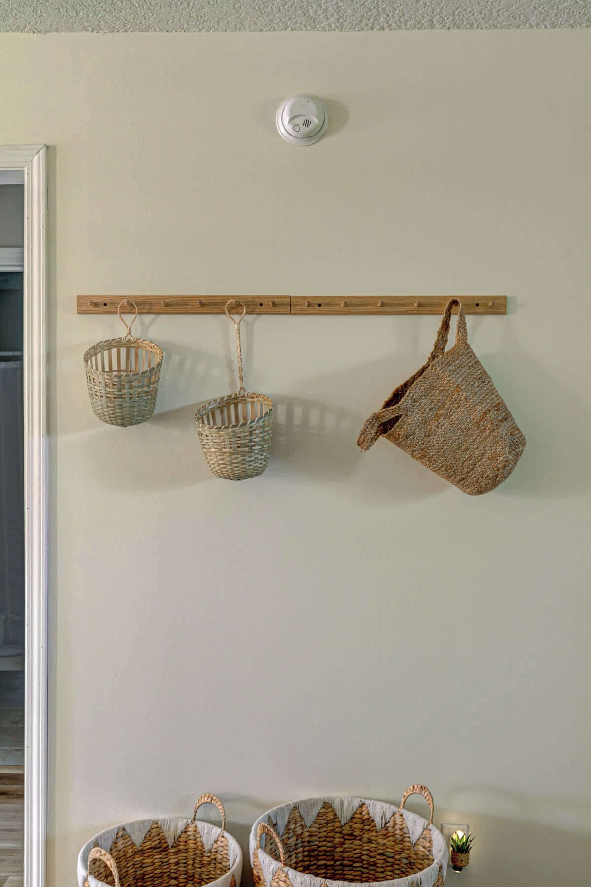 Un porte-objets en bois fixé au mur, contenant trois paniers tissés et un sac tissé.