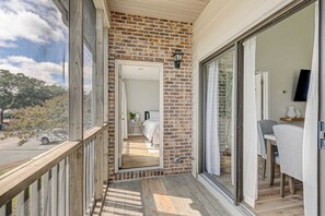Un balcon doté d’une balustrade en bois, d’un mur en briques et d’une porte-fenêtre coulissante donnant sur une chambre à coucher équipée d’un lit, d’une table de chevet et d’une plante.