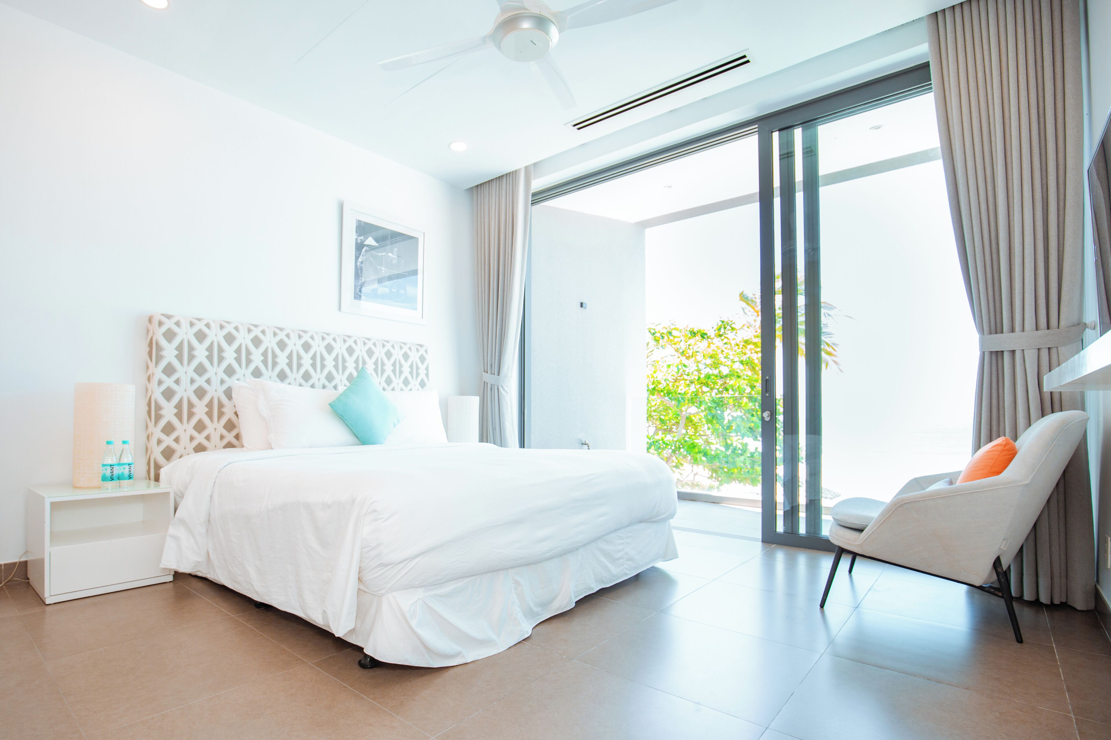Ein modernes Hotelzimmer mit einem großen Bett, einem weißen Nachttisch, einem Sessel und Blick ins Grüne durch die Glastür.