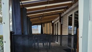 A covered outdoor seating area with a wooden deck and a black cushioned table. - Mobilhome saint hilaire de riez in 4 star campsite (Saint-Hilaire-de-Riez)