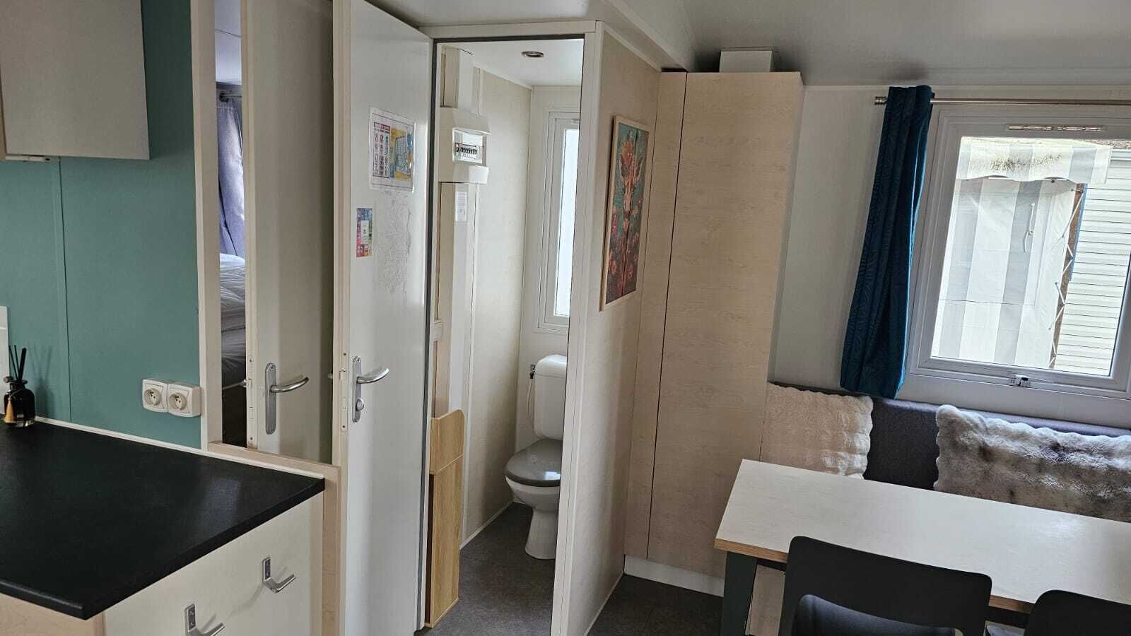 Een compacte kitchenette met een zwart aanrecht en een witte wastafel. Een klein zithoekje met een witte tafel en twee stoelen bevindt zich naast een raam met een blauw gordijn. In de achtergrond is een toilet zichtbaar.