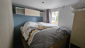 A small, narrow room with a single bed, a window with curtains, and a wall-mounted shelf. - Mobilhome saint hilaire de riez in 4 star campsite (Saint-Hilaire-de-Riez)