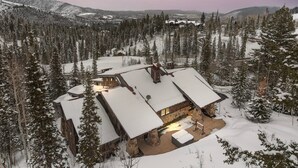 A snow-covered lodge with a stone exterior and multiple roofs, surrounded by dense forest and mountains. - Ski-in/ski-out luxe Colony chalet, views, hot tub, game room! (Park City)