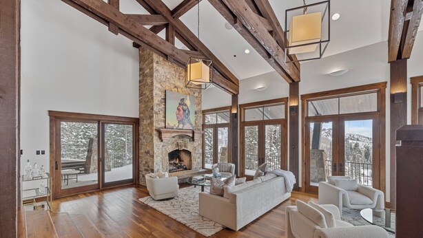 A spacious living room with a stone fireplace, wooden beams, large windows, and a cozy seating area. - Ski-in/ski-out luxe Colony chalet, views, hot tub, game room! (Park City)