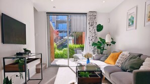 A modern living room with a grey sofa, a coffee table, a TV, and a sliding glass door leading to a balcony with plants.