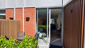 A modern building with a glass door leading to a well-lit interior, a wooden fence, and a small outdoor seating area with chairs and a table.