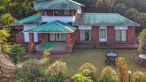 A two-story building with a green metal roof, brick walls, and a small front porch with a green mat. There is a patio area with outdoor furniture. - Amore Villa (Mahabaleshwar)
