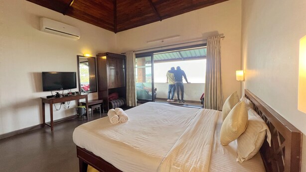 A hotel room with a large bed, a desk with a television, and a balcony with two people. - Amore Villa (Mahabaleshwar)