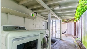 Un espace buanderie comprenant un lave-linge et un sèche-linge, une petite étagère avec des produits de nettoyage, et une vue sur une cour arrière avec une clôture en bois et une chaise.
