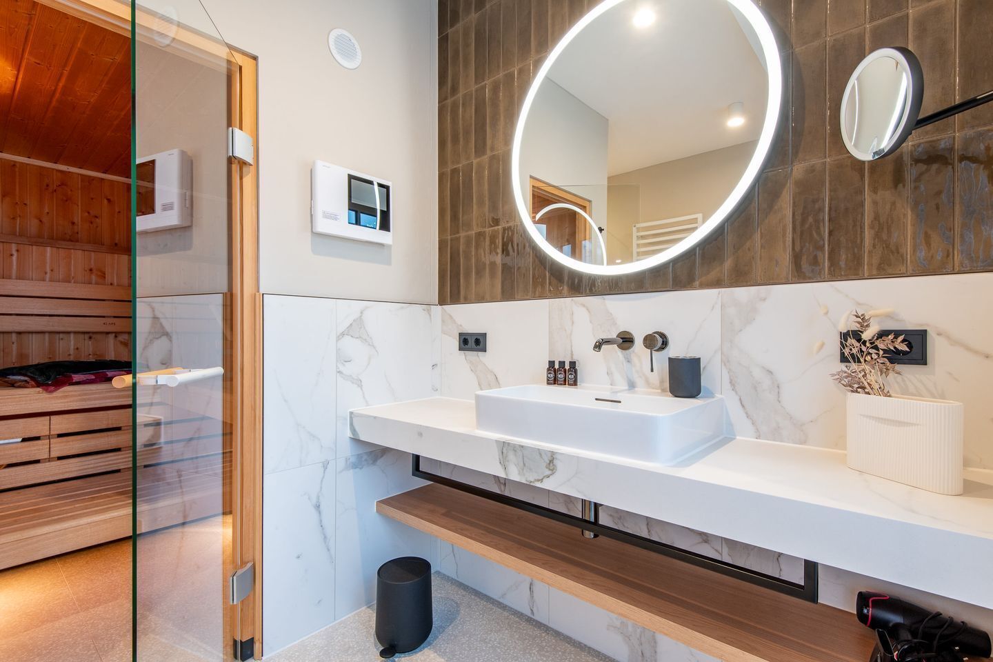 A modern bathroom with a wooden sauna, a white marble sink, and a large mirror with a circular light.