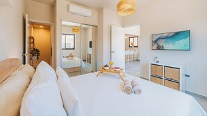 A modern hotel room with a large bed, a dining area with a table and chairs, a TV mounted on the wall, and a large window with a view of the beach.