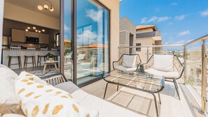 A balcony with a glass door leading to a kitchen, a sofa with patterned cushions, and two wicker chairs.