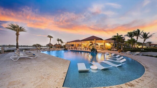 A resort pool with lounge chairs, palm trees, and a building with a red roof.