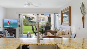 Cocina moderna con encimera de granito y pileta de acero inoxidable, un televisor mostrando un velero y vista a un patio con mobiliario exterior.