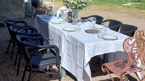 Un espacio de comedor al aire libre con una mesa preparada para cuatro personas, rodeada de sillas de mimbre, con un edificio de piedra y un campo arbolado de fondo.