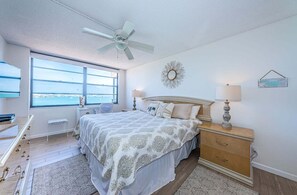 Une chambre à coucher comprenant un lit, des tables de chevet, un ventilateur de plafond, une fenêtre avec vue et un miroir décoratif.