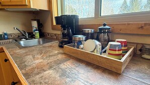 A kitchen with wooden cabinets, a coffee maker, a sink, and a window with a view of trees. - Alpine Horn #2 ~ Sauna & Close to Powderhorn! (Bessemer)