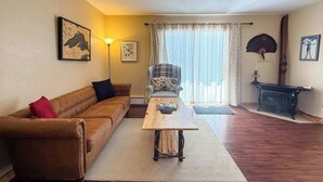 A living room with a brown sofa, a wooden coffee table, a patterned armchair, a fireplace, and a large window with curtains. - Alpine Horn #2 ~ Sauna & Close to Powderhorn! (Bessemer)