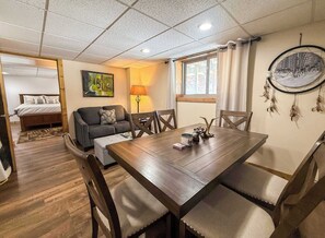 A room with a wooden dining table, chairs, a sofa, a bed, and a lamp.