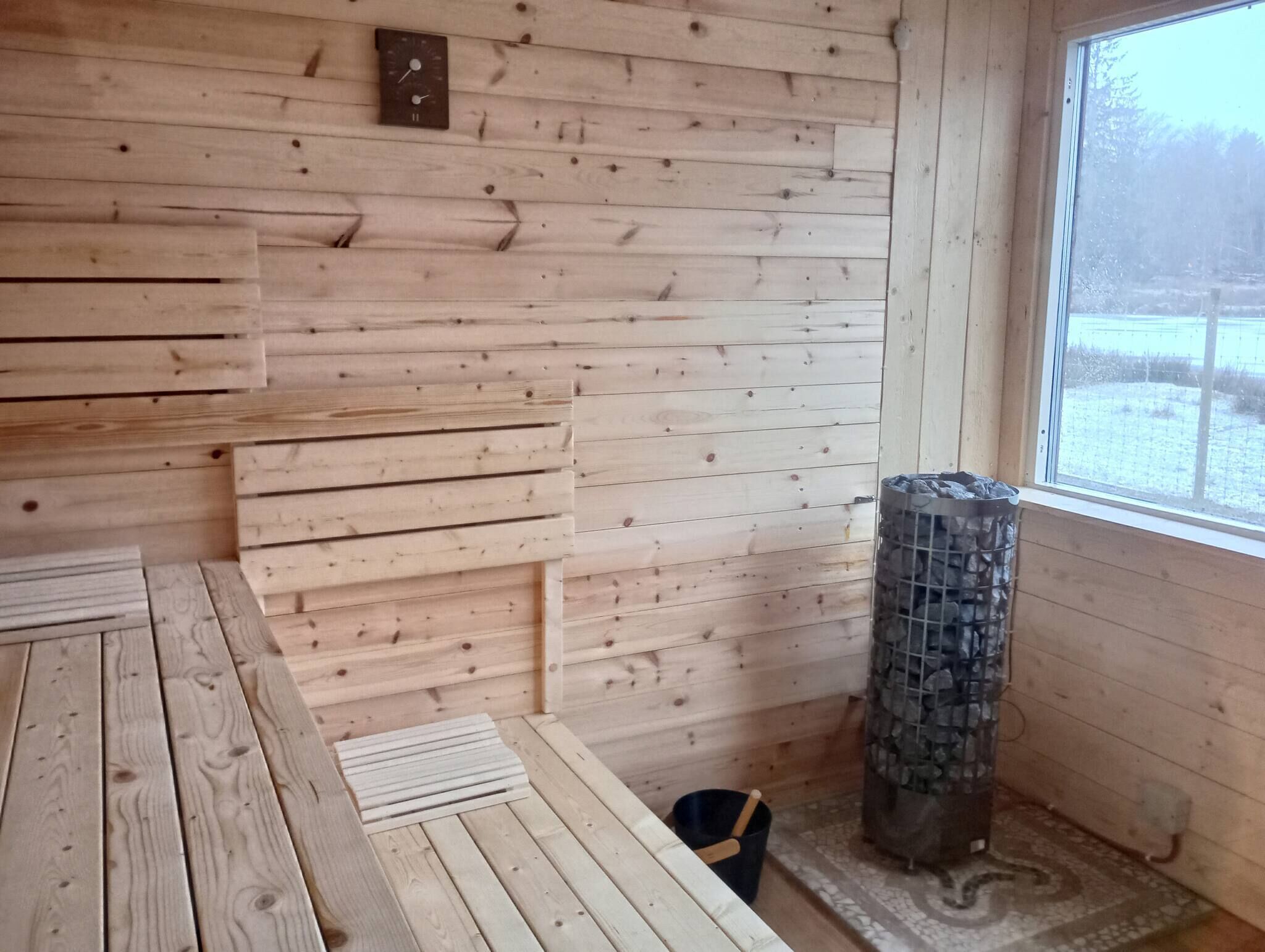 A wooden interior with a wooden bench, a stone heater, and a window with a view of a snowy landscape.