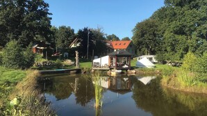 A serene lakeside setting with a small house featuring a red roof, surrounded by lush greenery and a calm pond reflecting the sky. - Viltpark Rollerbü Hjortaröd (Tyringe)