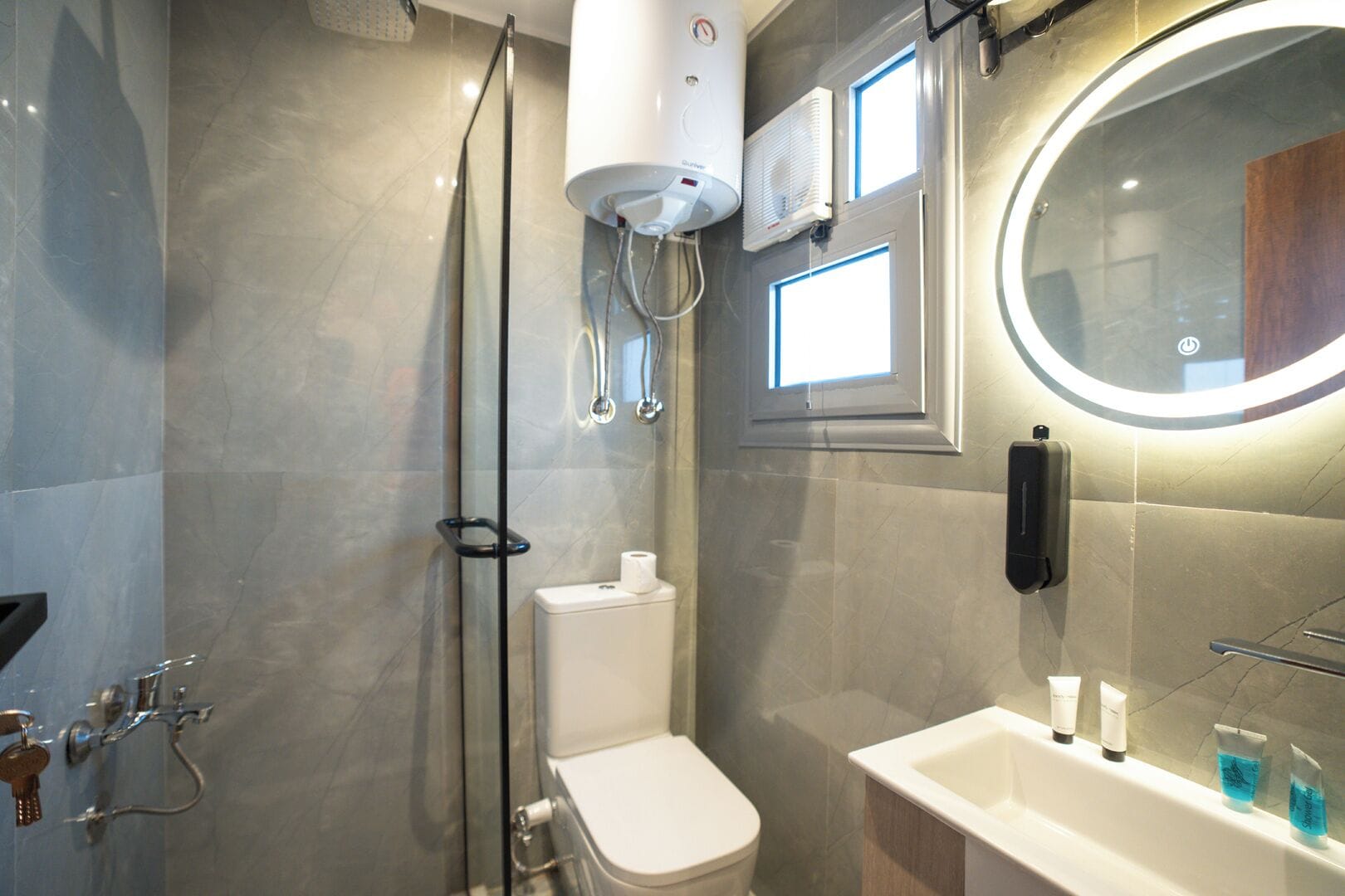 A modern bathroom with a glass-enclosed shower, a toilet, a sink with toiletries, and a mirror with a light.