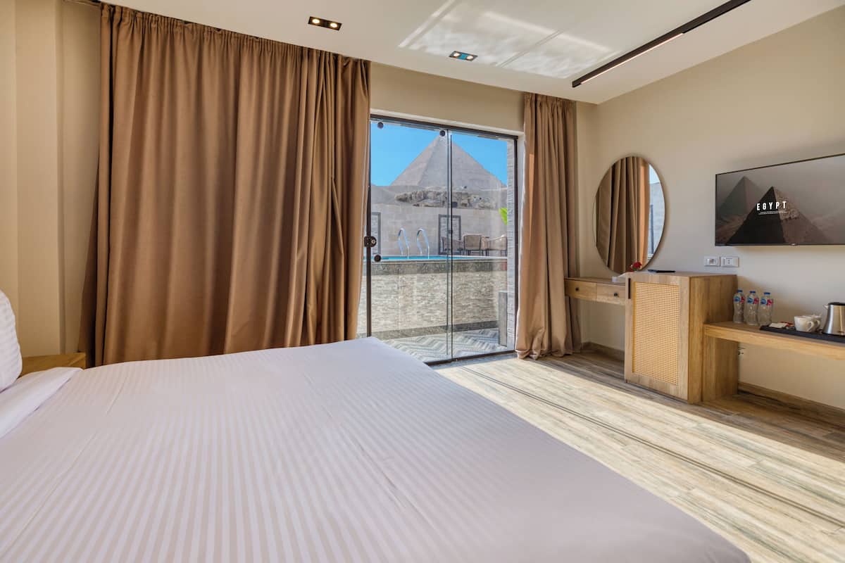 Habitació d'hotel amb un llit gran, una porta corredissa de vidre que dona a un balcó amb vista a una piràmide, un escriptori de fusta i un televisor penjat a la paret.