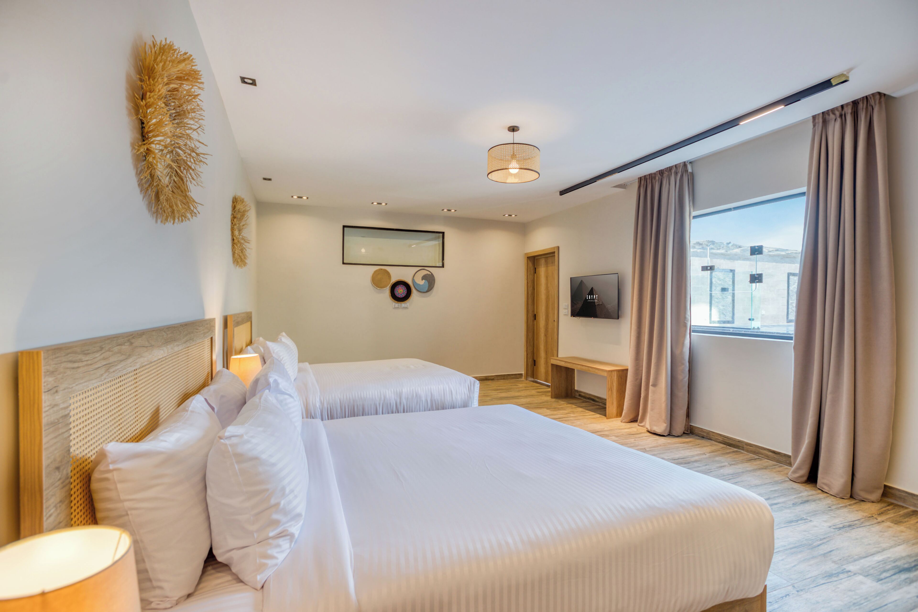 Ein Hotelzimmer mit zwei Betten, einem Fernseher, einem Schreibtisch und einem großen Fenster mit Vorhängen.