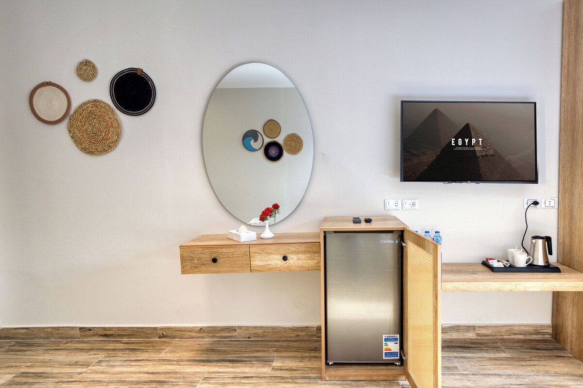 Una habitación de hotel moderna con un escritorio de madera, un refrigerador pequeño, un espejo y un televisor de pantalla plana mostrando 'EGYPT'.