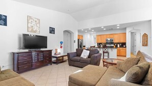 A spacious living room with a flat-screen TV, a sofa, and a coffee table. A kitchen with wooden cabinets and a dining area is visible in the background. - Family Friendly  w/ Badminton | Goodyear Park (Goodyear)