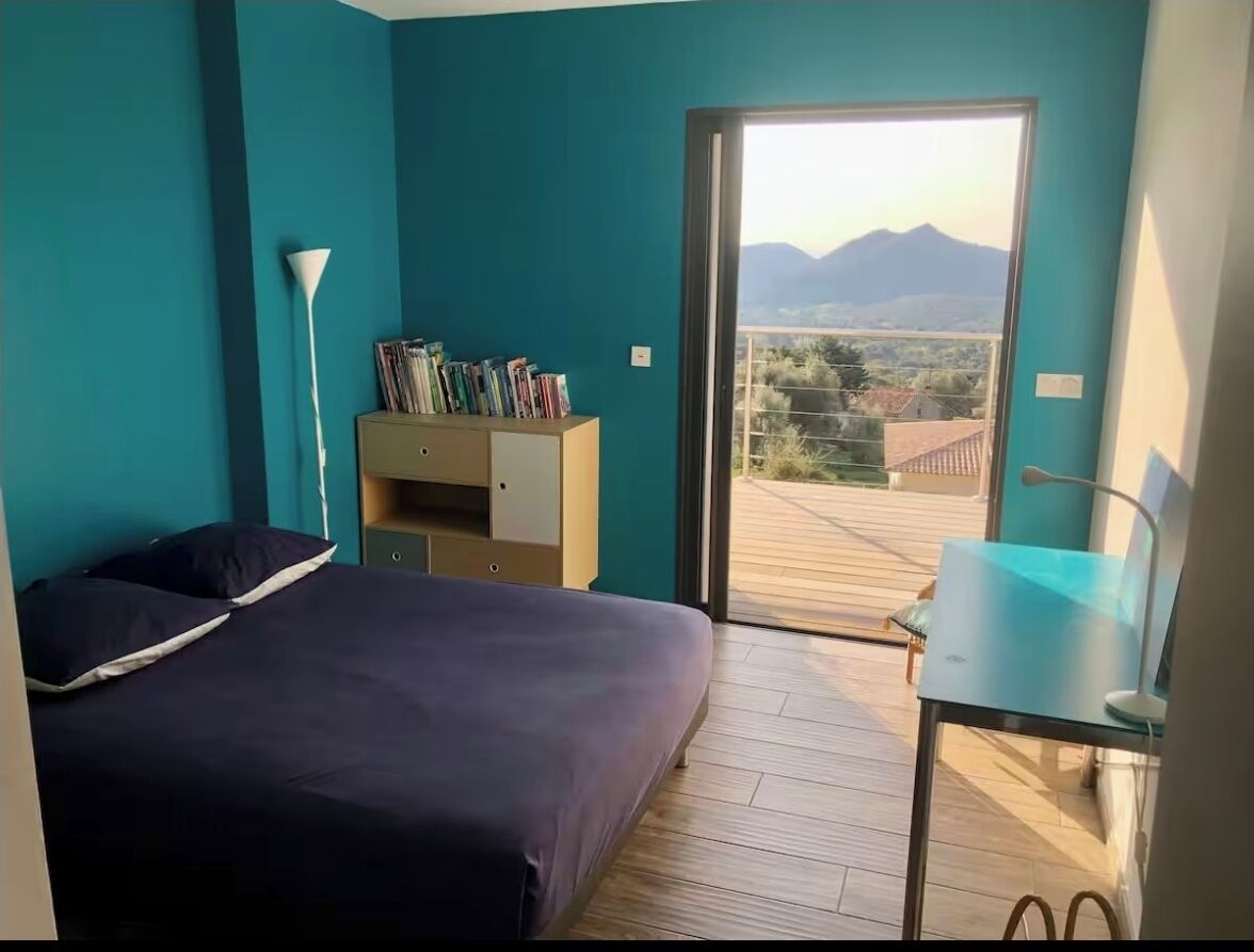 A bedroom with a bed, a desk, a bookshelf, and a view of the outdoors.
