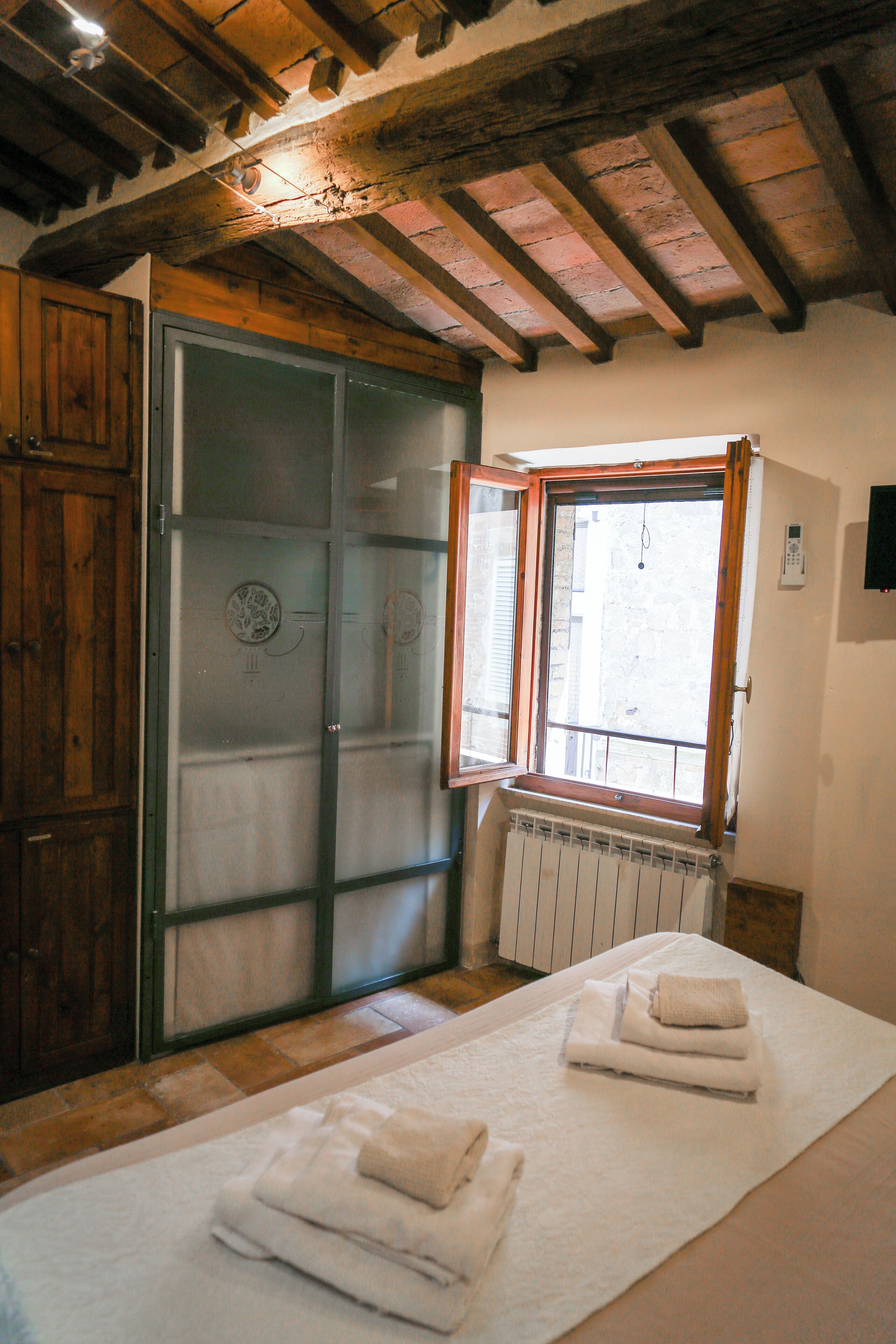 Una stanza con soffitto in legno, un'ampia finestra con cornice in legno, un termosifone sotto la finestra e un letto con asciugamani piegati su un tappeto bianco.