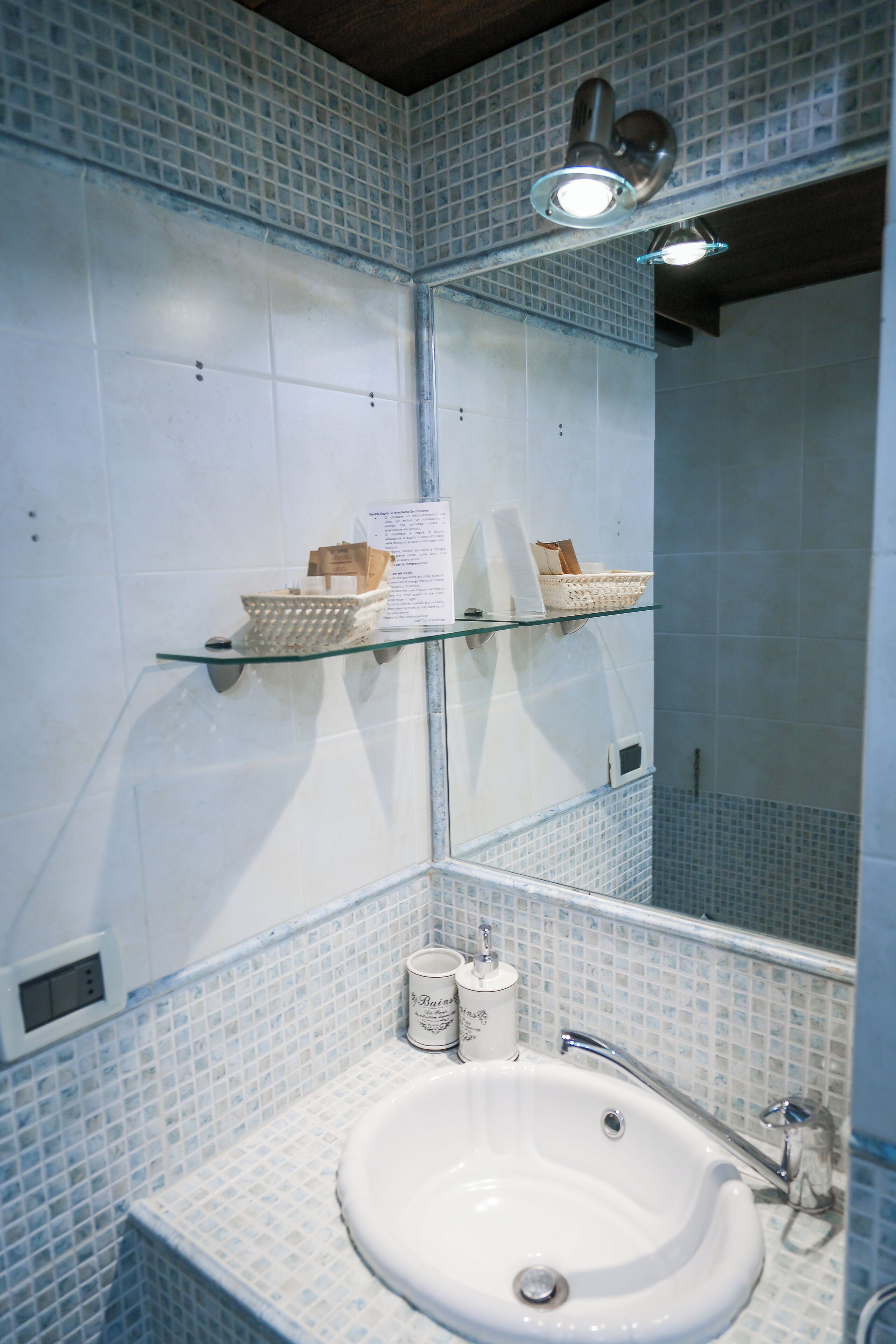 Une salle de bain avec un lavabo blanc, une étagère en verre où sont rangés les articles de toilette, et un miroir au-dessus du lavabo.