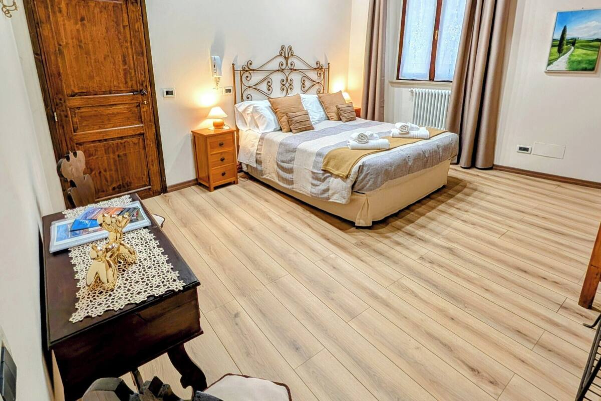 A bedroom with a wooden floor, a bed with a headboard, a desk with a chair, a wooden door, and a window with curtains.