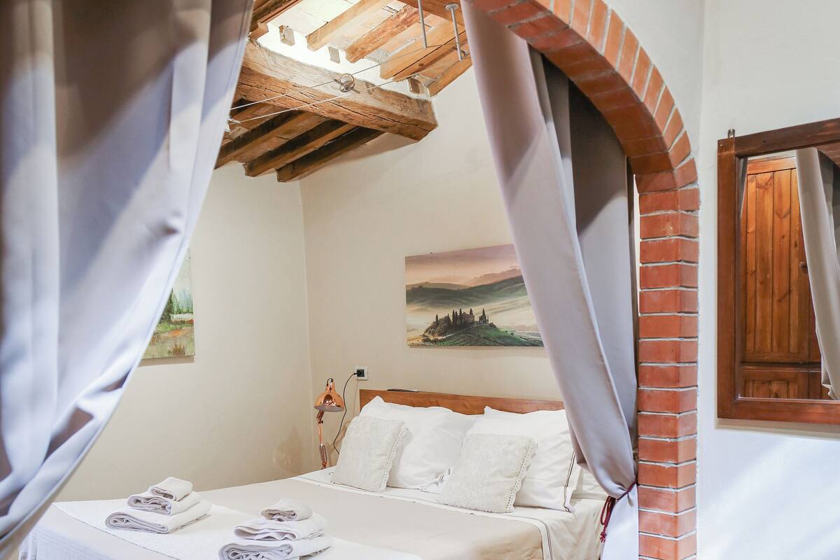 A bedroom with a brick archway, a bed with white linens, a small table with a magazine, and a painting on the wall.