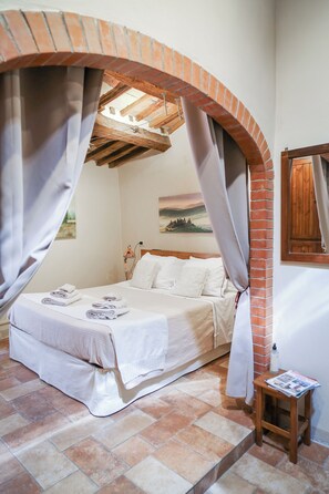 A bedroom with a brick archway, a bed with white linens, a small table with a magazine, and a painting on the wall. - TuscanyLettings Studio&Room (Pienza)