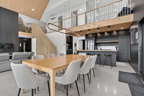 A modern interior with a wooden dining table, grey upholstered chairs, a kitchen area, and a staircase leading to an upper level.