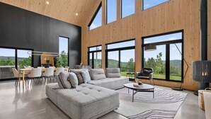 A modern living room with a sectional sofa, a dining area with a table and chairs, large windows offering a view of the outdoors, and a fireplace.