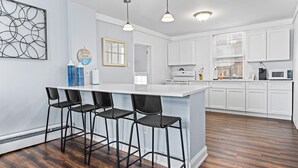 A modern kitchen with white cabinets, a central island with bar stools, pendant lighting, and a wooden floor.