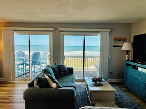 A living room with a sofa, a coffee table, and a TV. There are two sliding glass doors leading to a balcony with beach chairs and a view of the ocean. - Oceanfront 2 bedroom condo with pool in Carolina Beach (Carolina Beach)