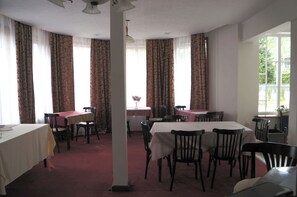 A dining area with tables and chairs, large windows with curtains, and a view of greenery outside.