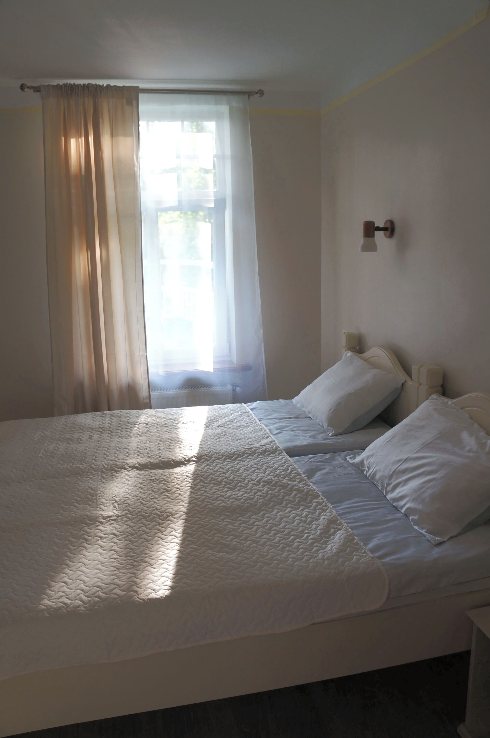 Una cama con ropa blanca, una ventana con cortinas traslúcidas y una lámpara fijada a la pared.