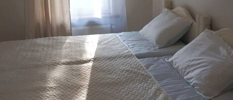 Una cama con ropa blanca, una ventana con cortinas traslúcidas y una lámpara fijada a la pared.